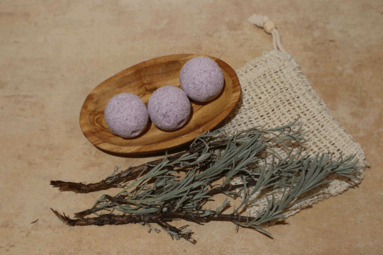 Dies ist ein Foto von einer violette Bio Badekugel Lavendel der Firma Mutschs mit einem Lavendelzweige und einem Seifensäckchen.